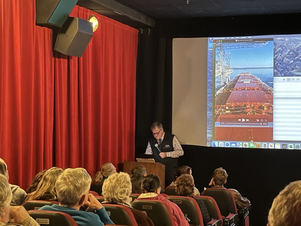 A man stands at a podium talking to an audience in a theater with a screen behind him.