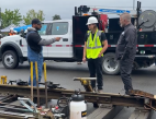 Two men at a rail track demonstration speak to a student in safety gear.