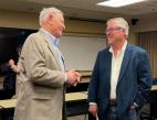 Two older men smile at each other at a public meeting.