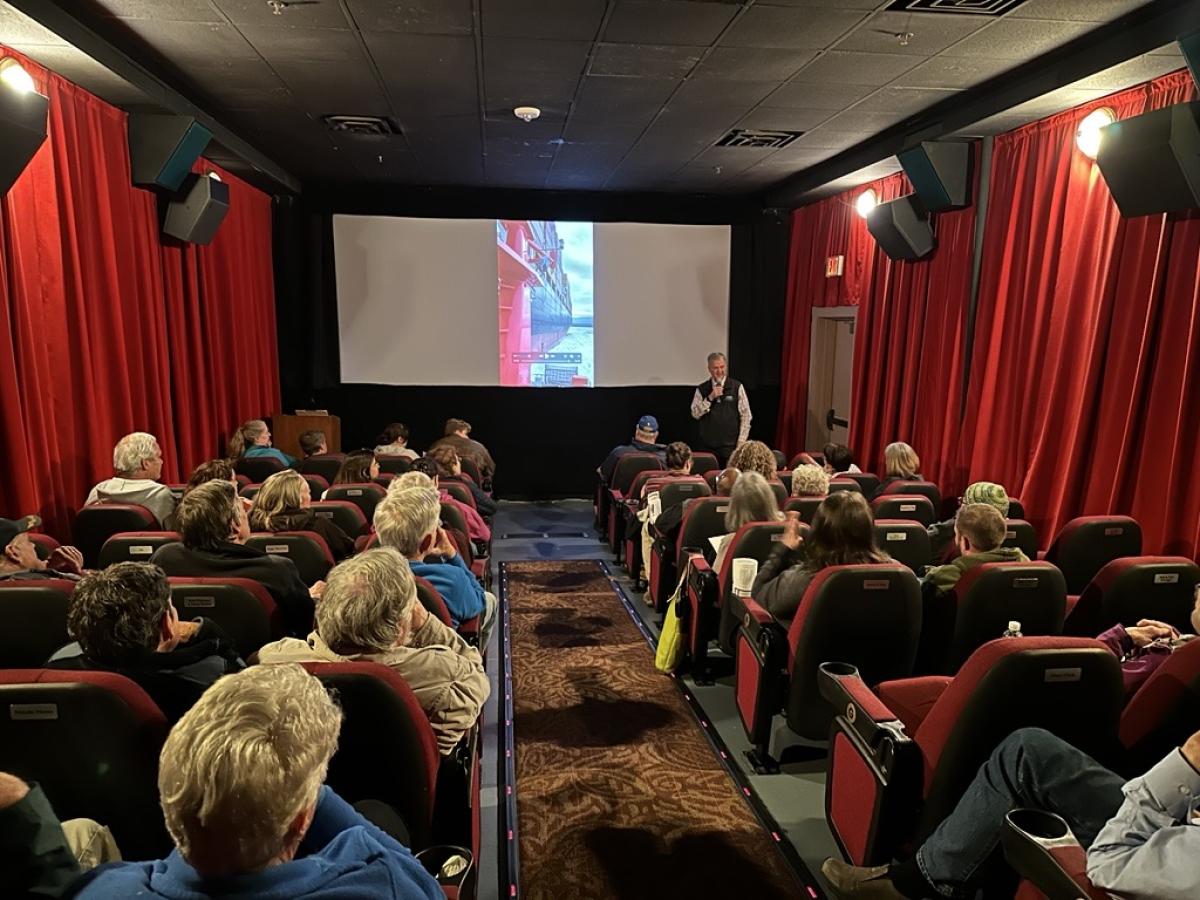 A group of people sit in chairs in a small theater while a man gives a presentation.