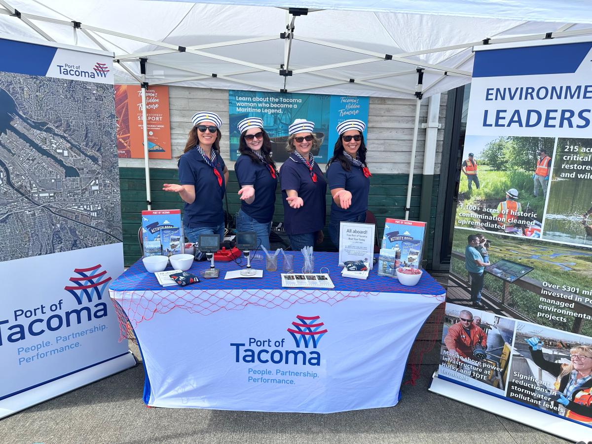 Four women in sailor hats hold their hands out while standing behind a Port booth at Oceanfest 2024.